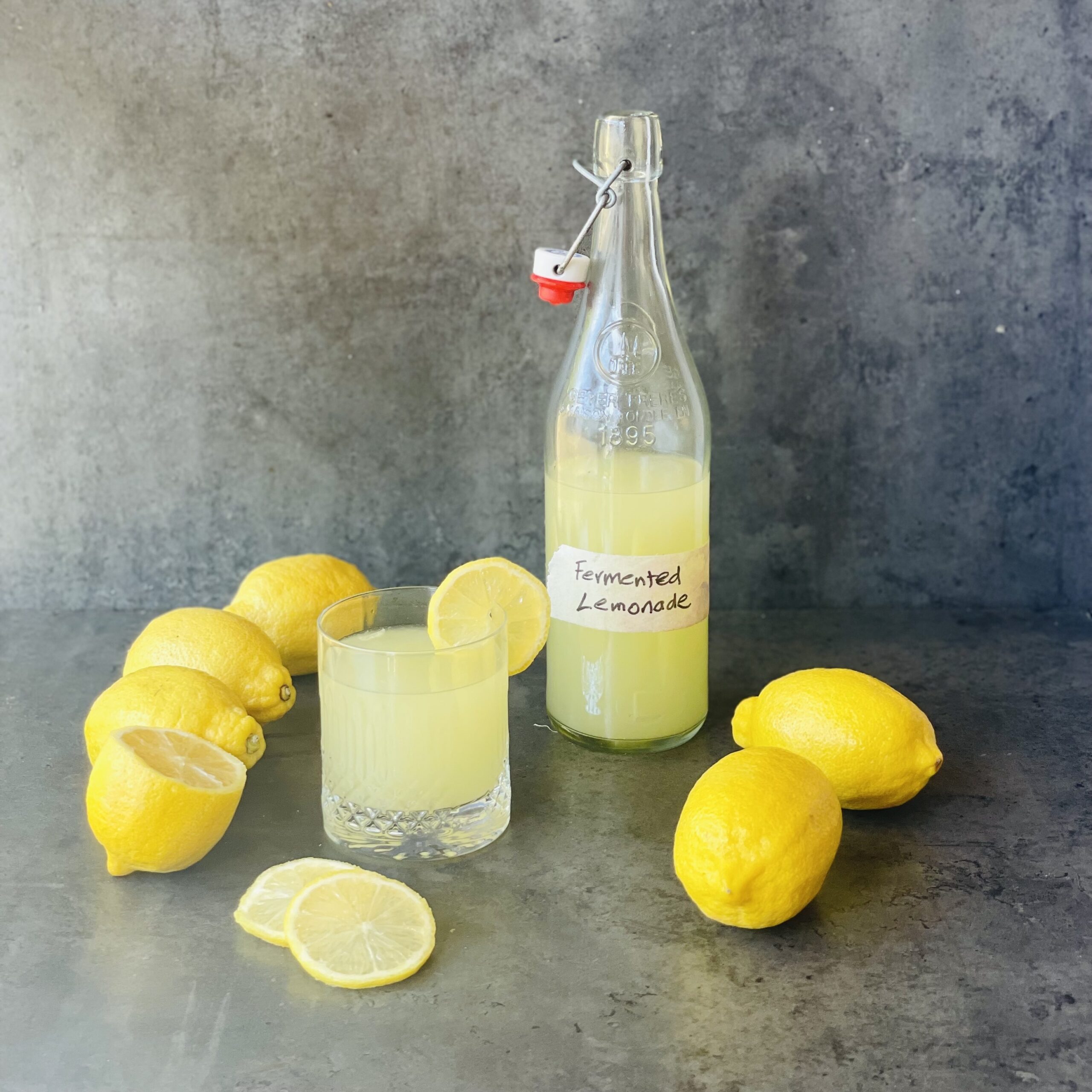 A photo of a glass bottle of fermented lemonade, a small glass of lemonade next to it and lemons